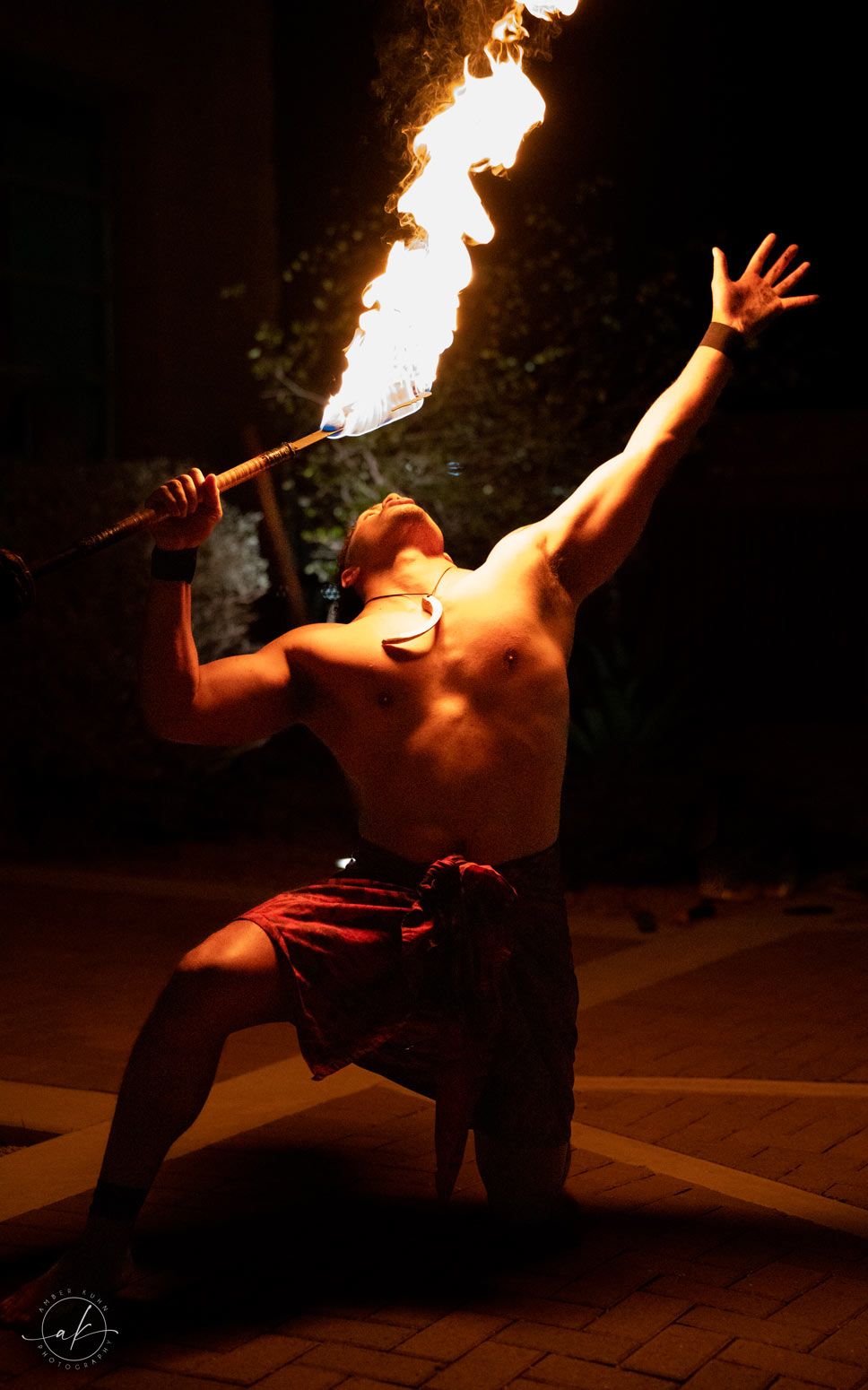 Fire performer in red shorts creates dramatic display with flaming torch against dark background.