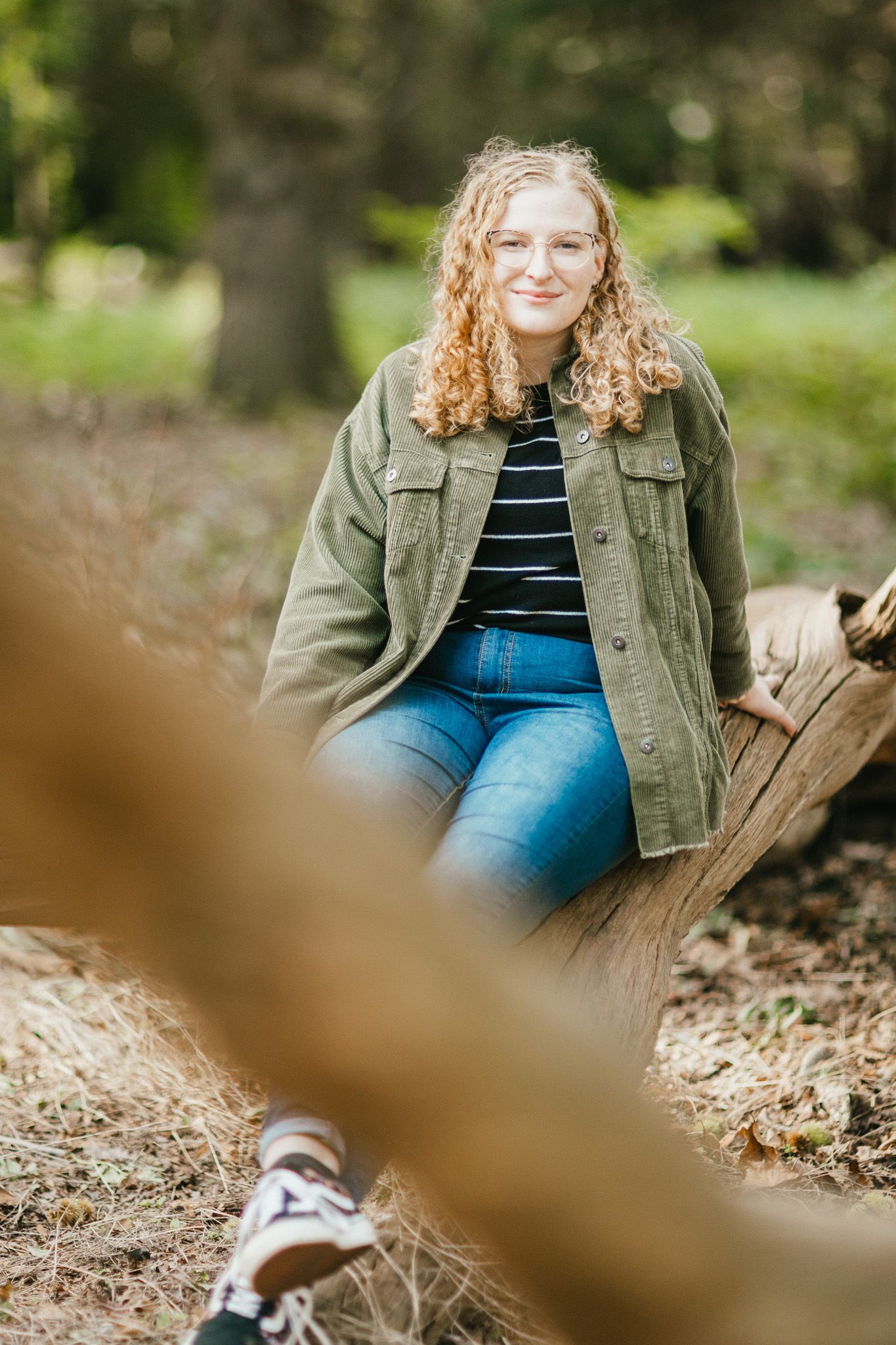 Woman in green jacket and striped shirt sitting on wooden log in outdoor nature setting.