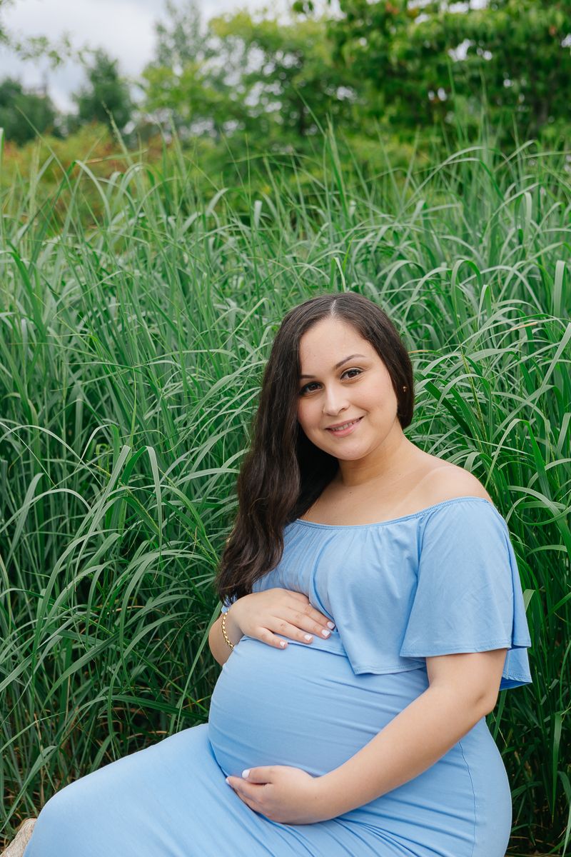A woman wearing a blue dress sits cradling her tummy against tall grass during a maternity photo shoot by Paula Ortiz NY
