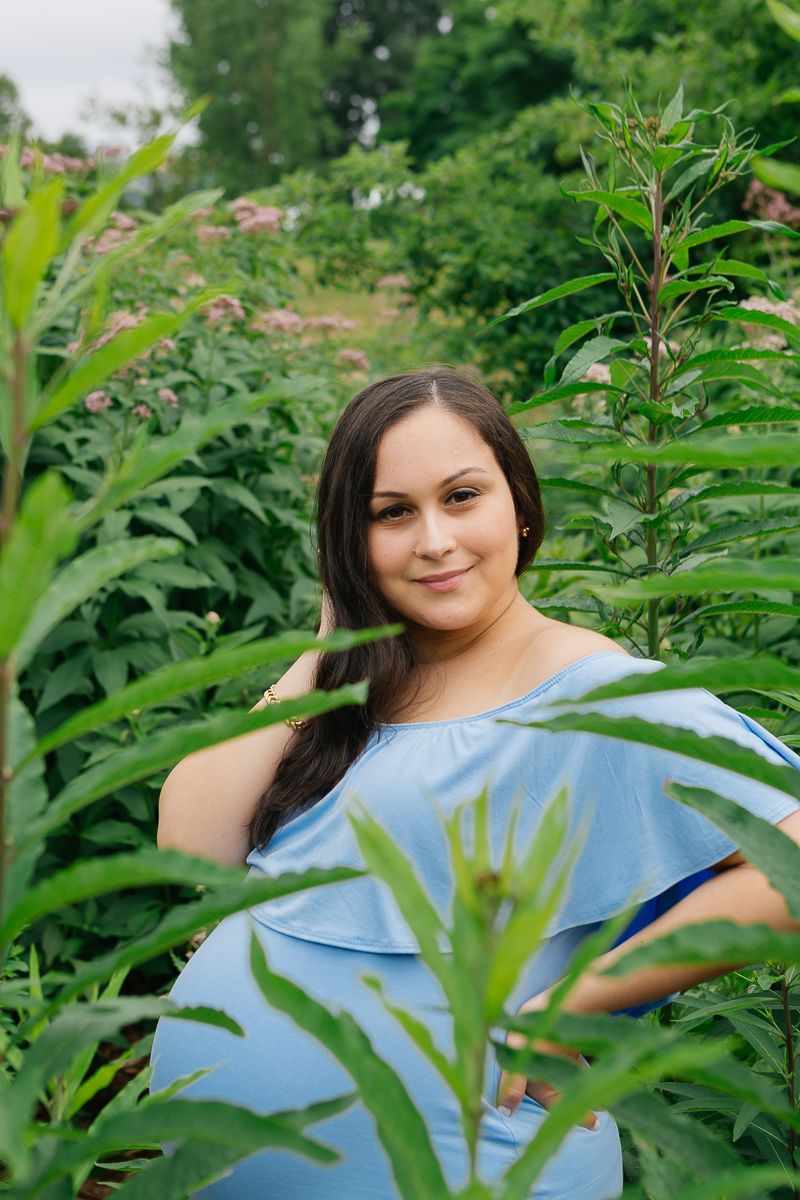 A pregnant woman in a blue dress stands amongst tall greenery, photo by Paula Ortiz NYC maternity photographer