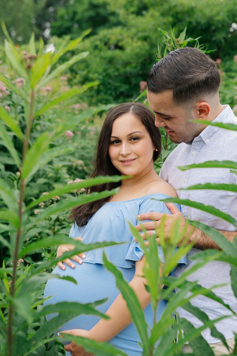 a couple stands together amongst tall greenery in Roosevelt Island during their maternity session