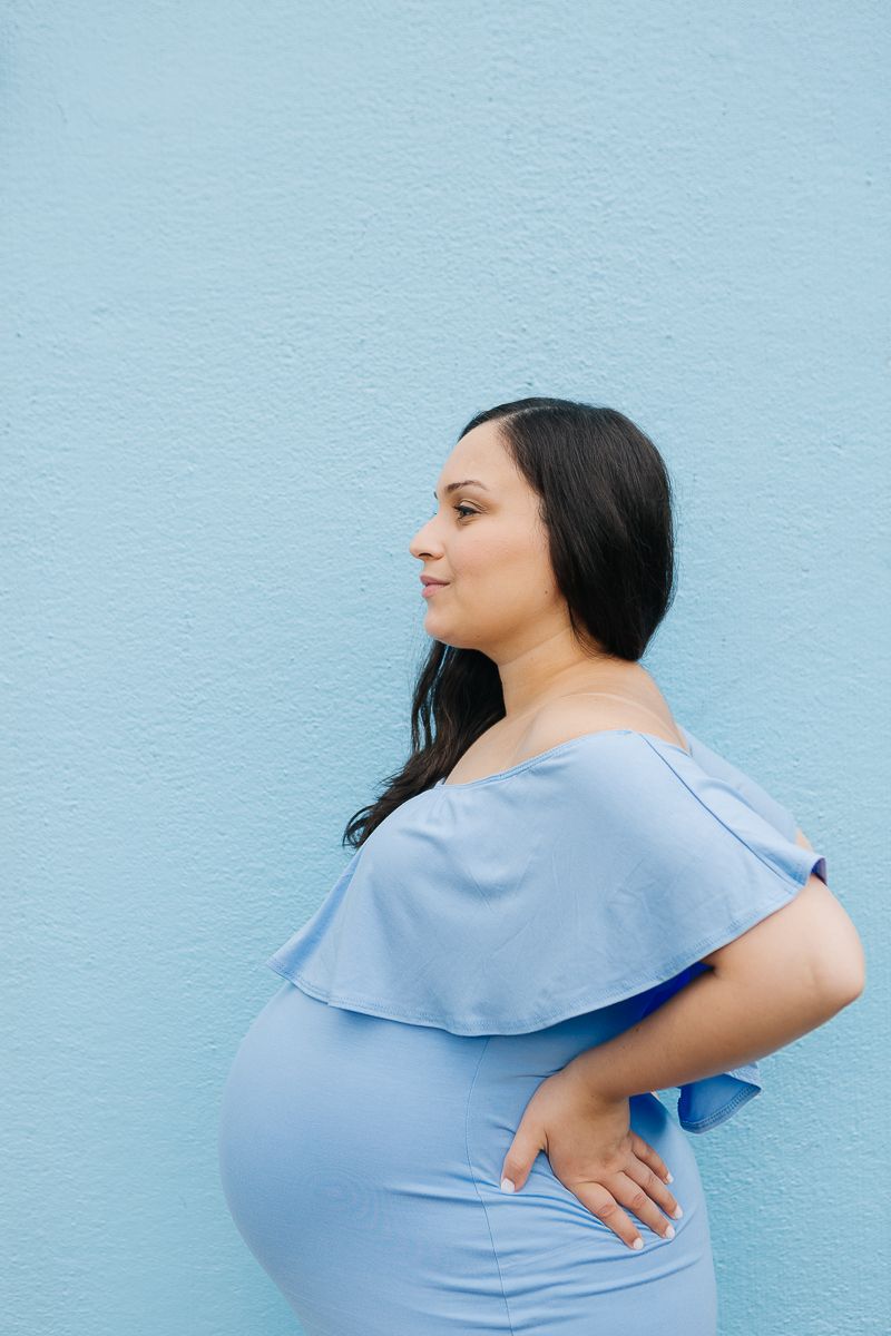 a profile view of a pregnant woman in a blue dress standing in front of a blue wall during her maternity session in NYC