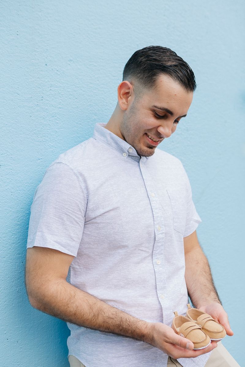 A dad to be leans against a blue wall while holding baby shoes during a maternity session by NYC maternity photographer