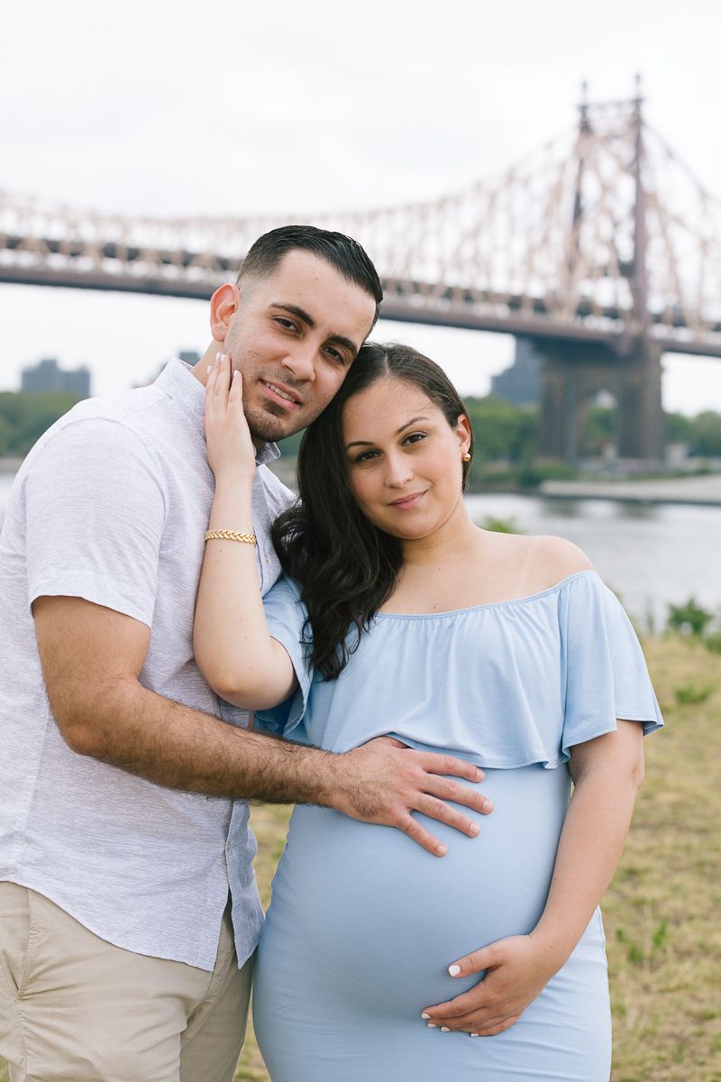 a couple stands next to each other with a NYC bridge in the background during a maternity session in Roosevelt Island