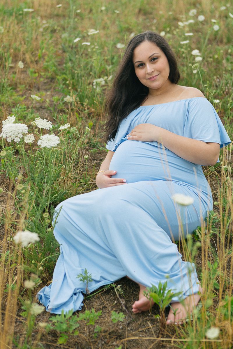 a pregnant woman in a blue dress lays on the grass amongst white flowers durning her maternity outdoor photo session NYC