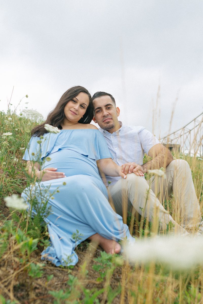 a couple sits amongst grass and flowers during their maternity session in Roosevelt Island NYC by Paula Ortiz NYC family