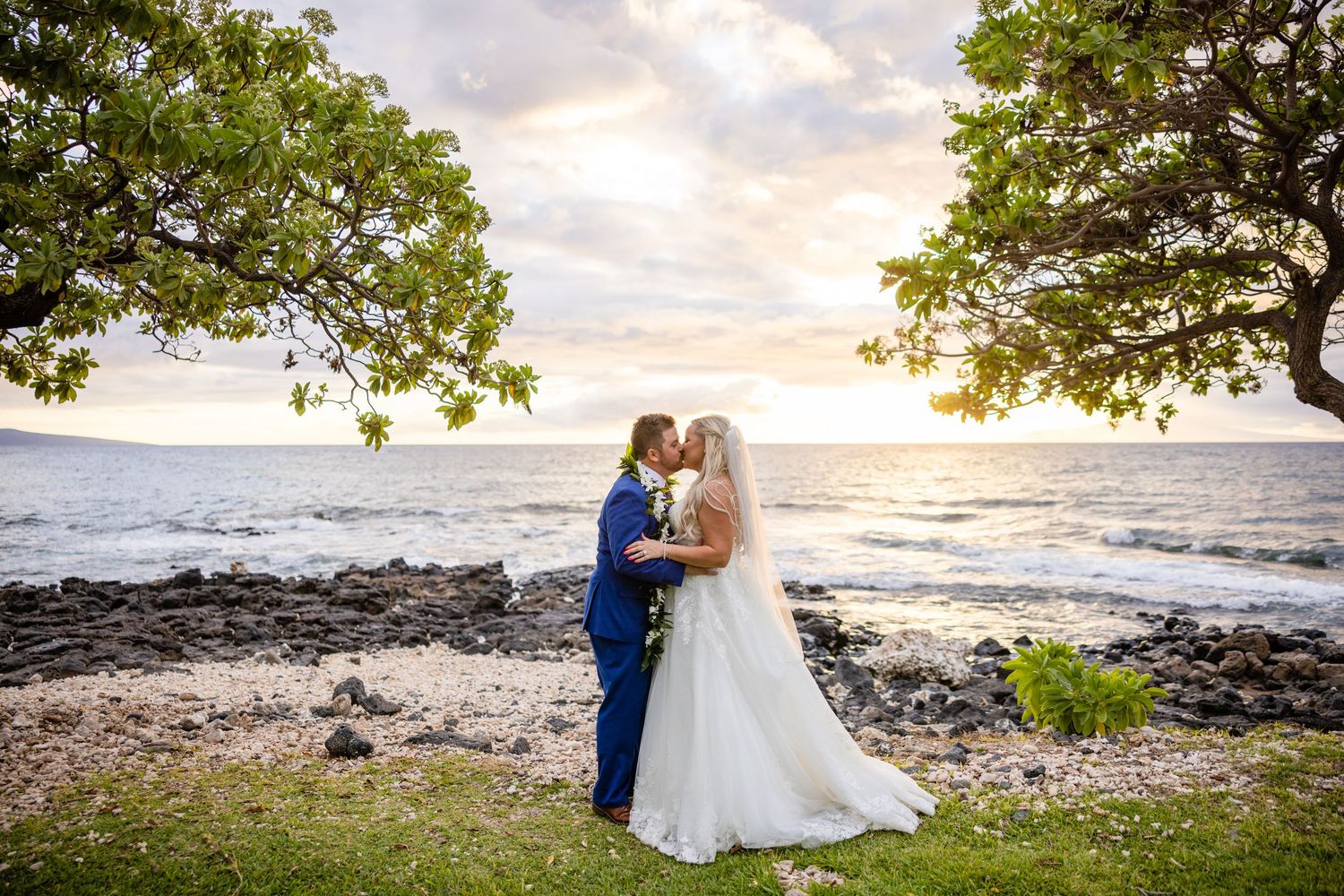 Couple shares sunset kiss by rocky oceanside shore under lush green trees.