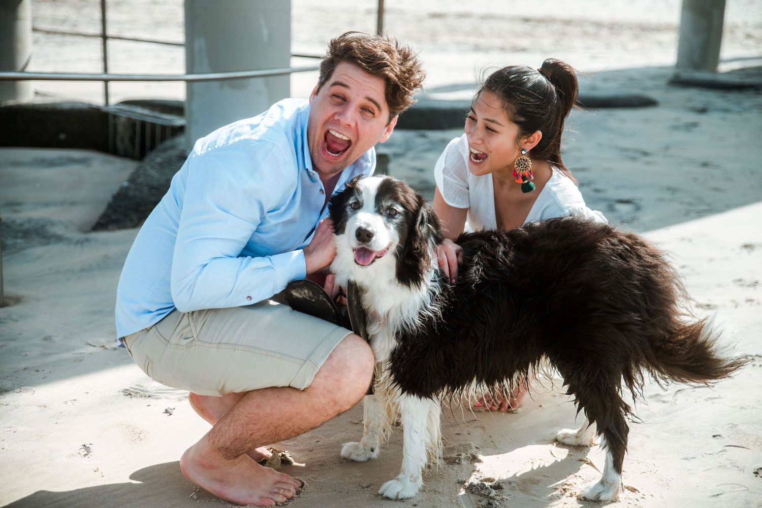 A fun beach photoshoot with a happy Border Collie dog and two people crouching down under a pier.