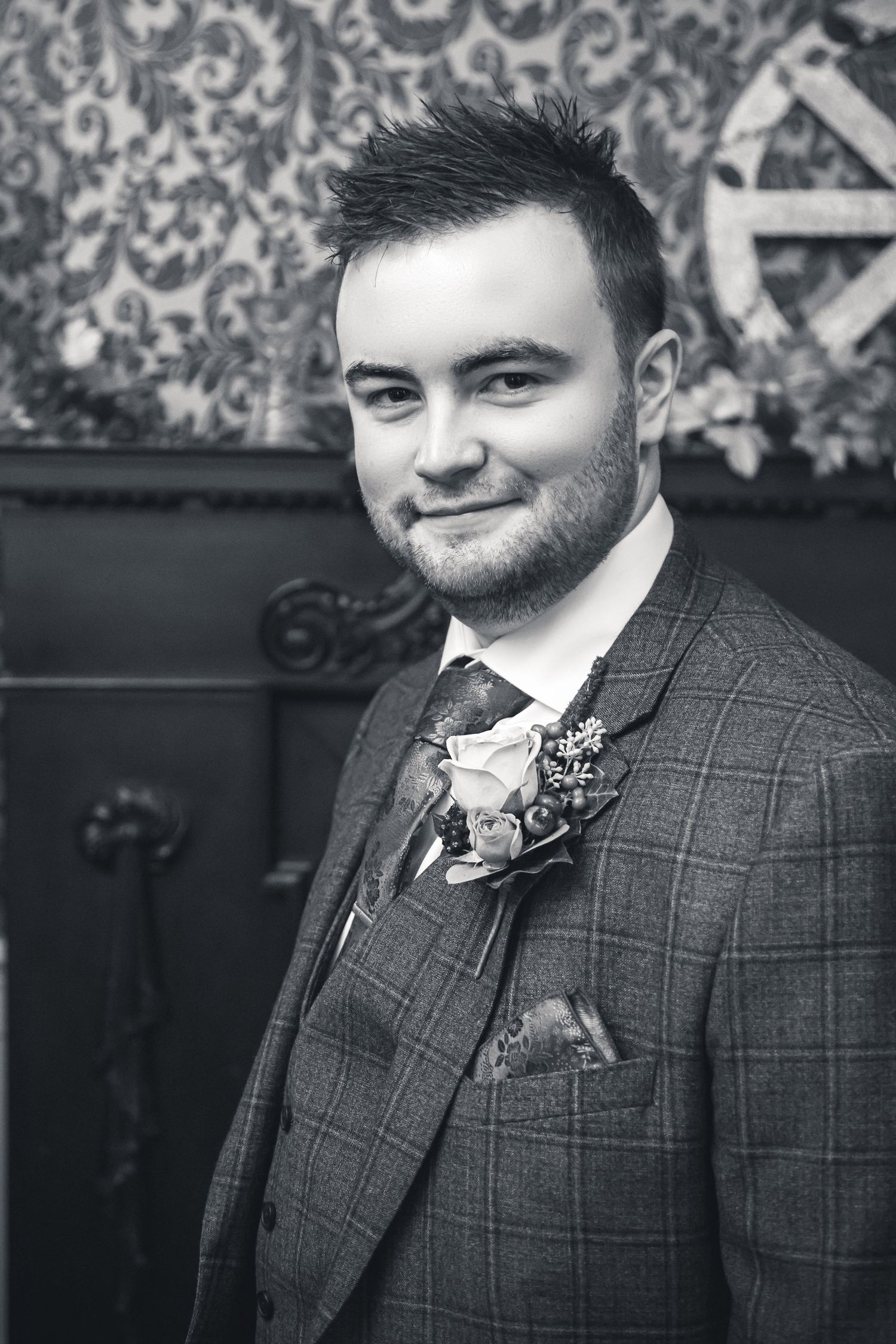 A groom in a checkered suit and burgundy tie smiles at camera against patterned wallpaper.