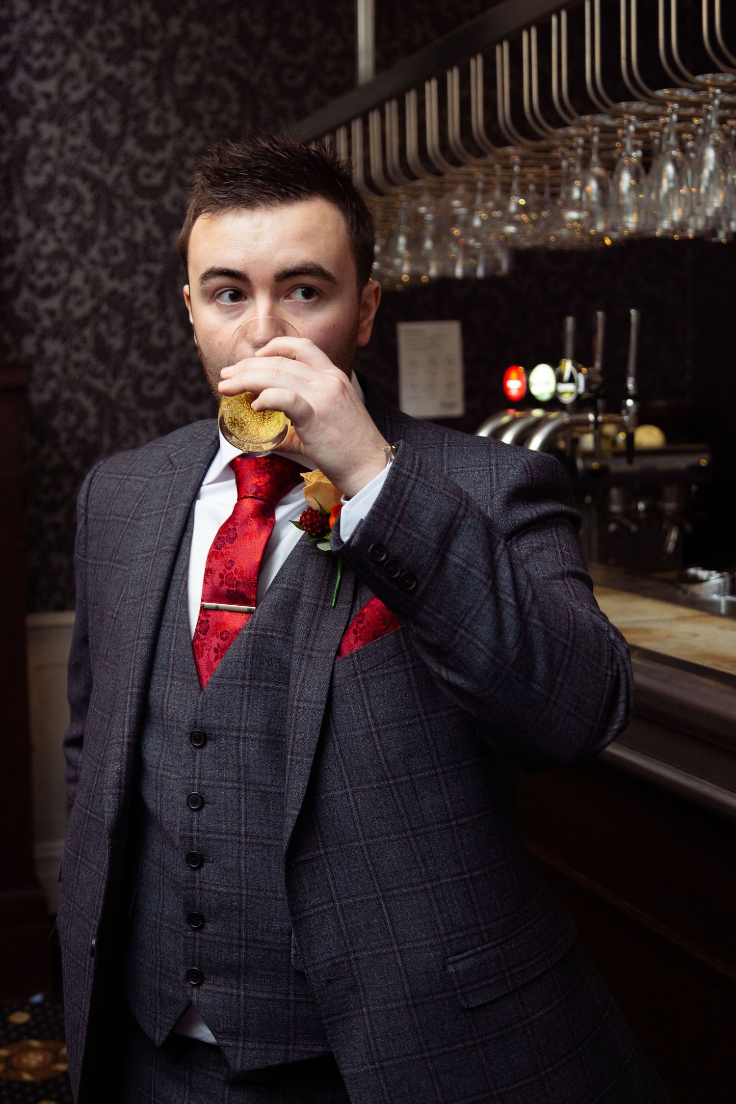 A groom in a three-piece suit takes a drink at a bar with hanging glasses visible.