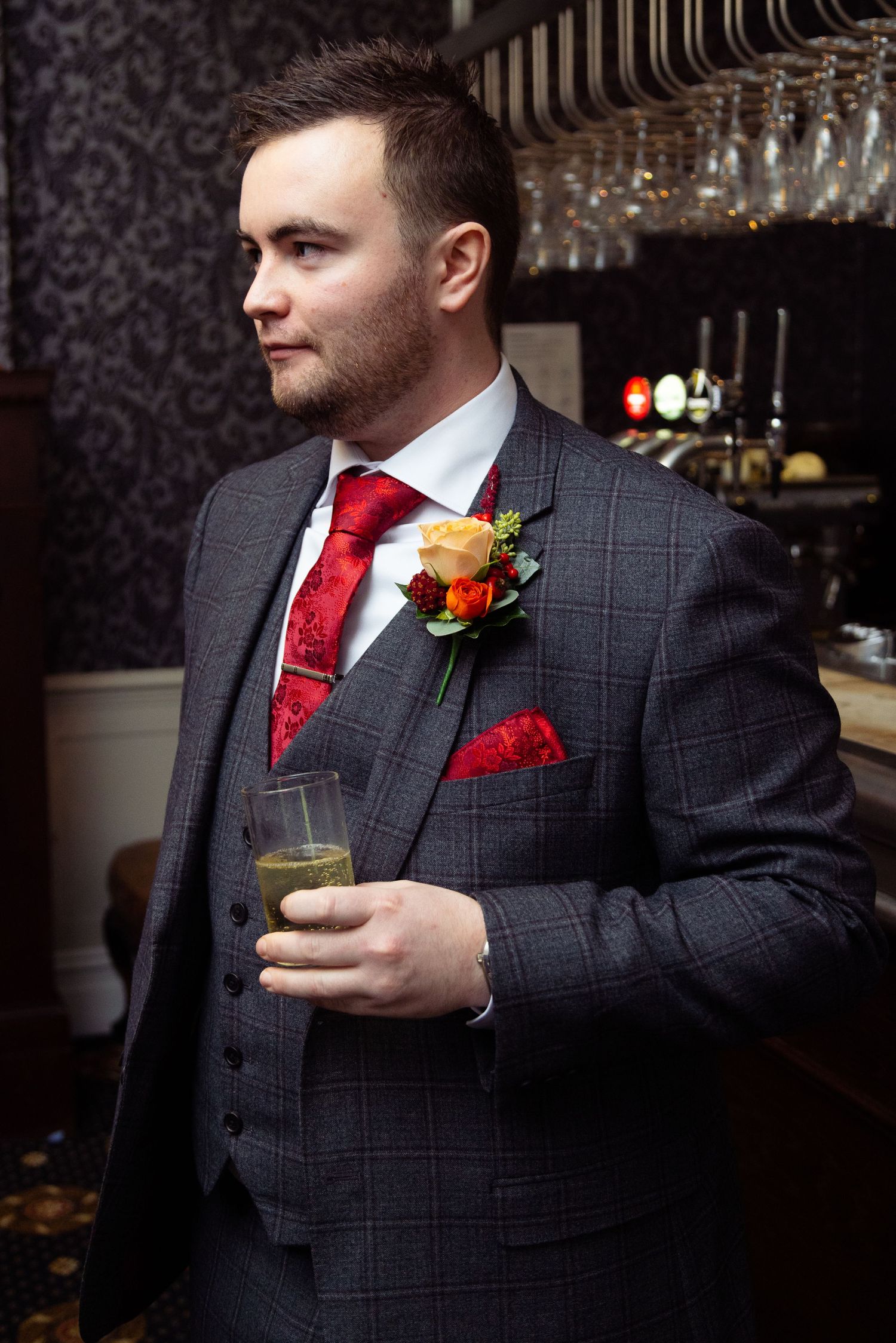 A groom in gray checkered suit with red tie poses with drink against bar backdrop.