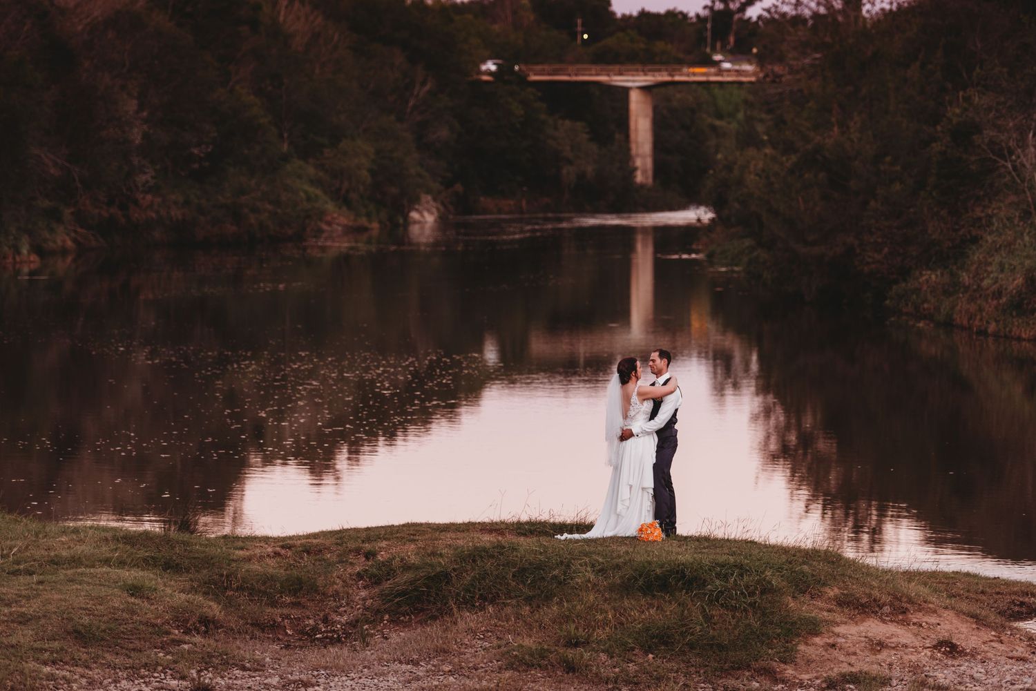 Couple standing by lake at dusk with bridge reflection in water.