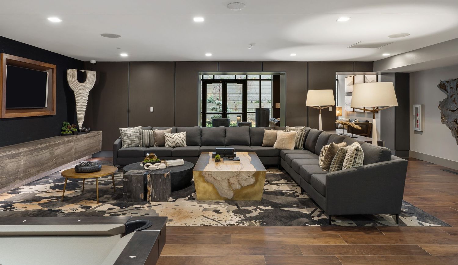 Modern apartment community room with dark sectional sofa, gold accent tables and cowhide rug on wood flooring.