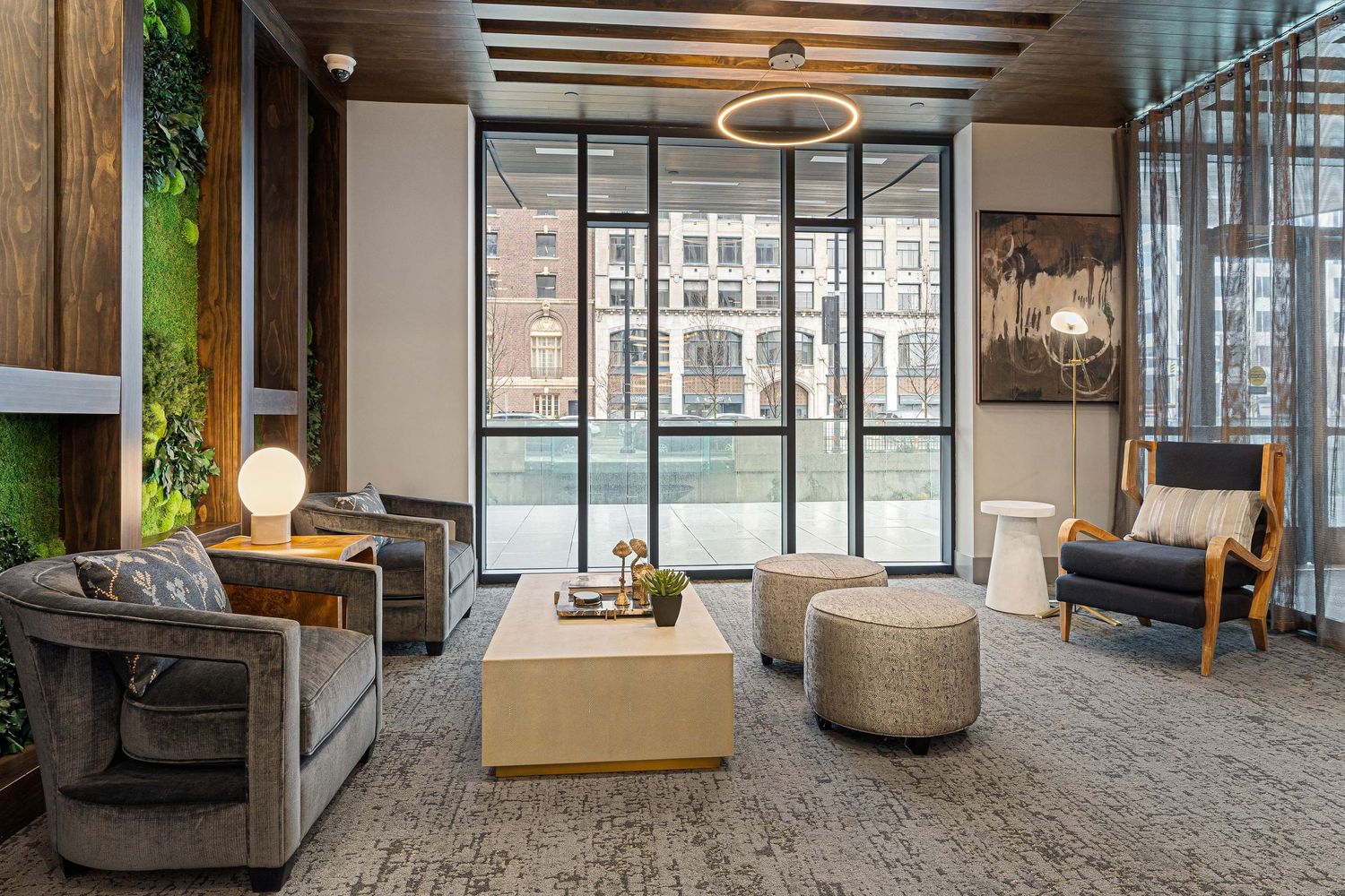 Modern apartment lobby with grey seating, wooden accents, and floor-to-ceiling windows overlooking a city view.