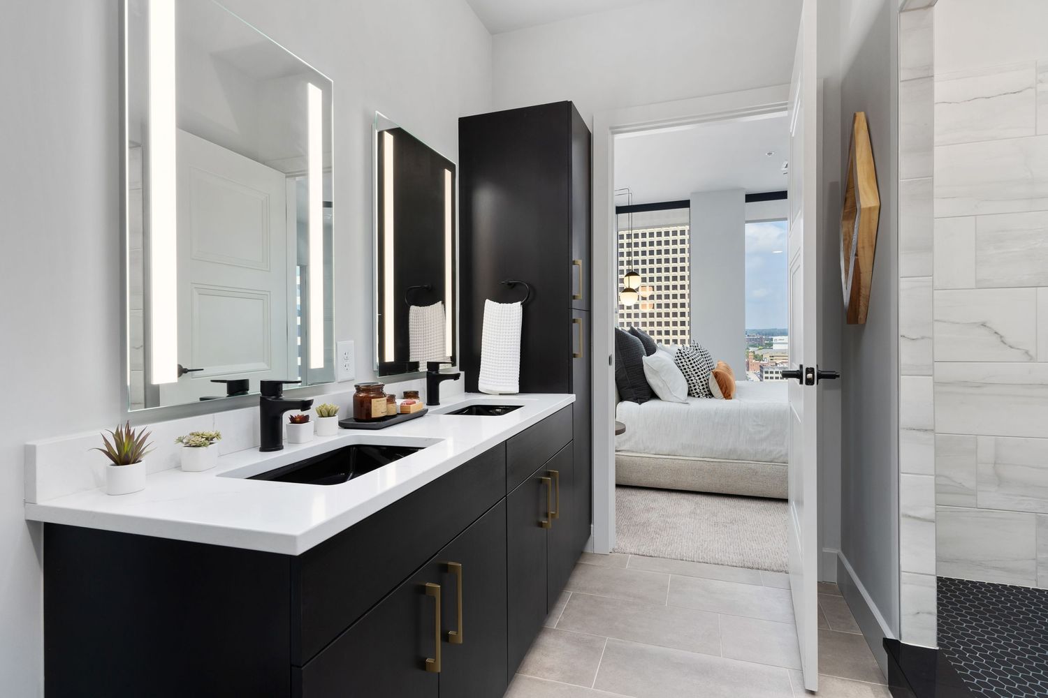 Modern apartment bathroom features black vanity cabinet with white countertop, dual sinks, and matte bronze hardware leading to bedroom.