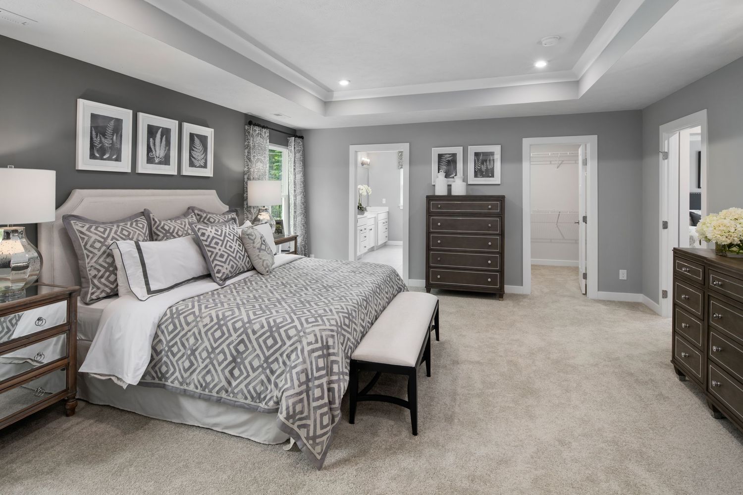 Luxurious primary bedroom with gray and white decor, tray ceiling, plush carpet and dark wood furniture in a model home.
