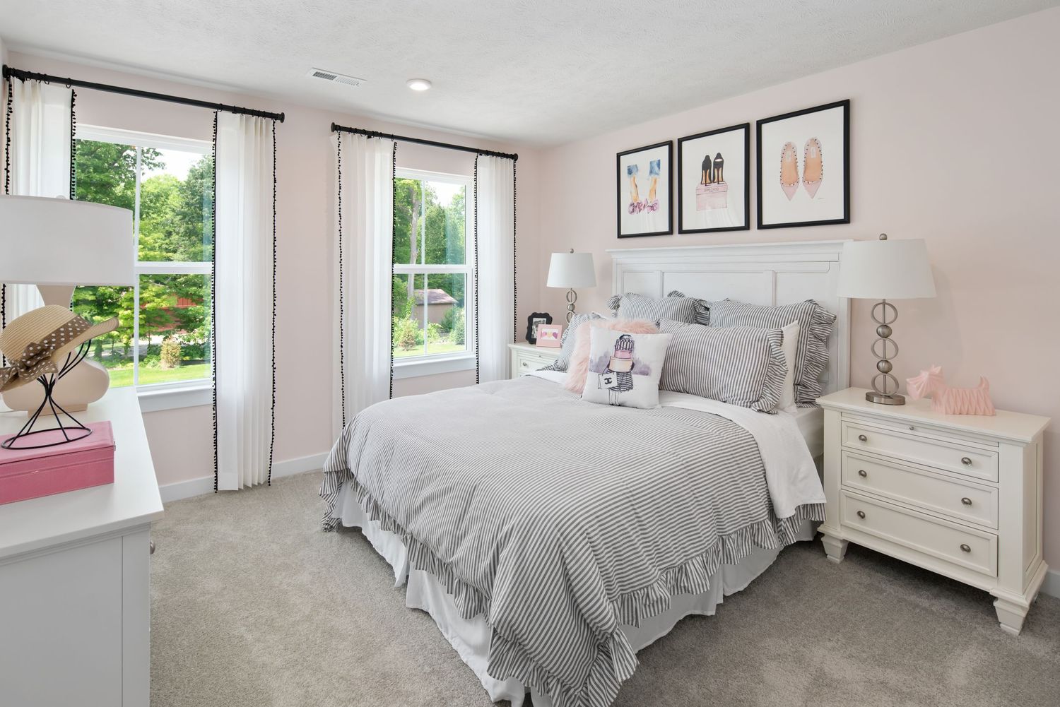 A bright and cozy model home bedroom with gray bedding, white furniture, framed wall art, and natural light from large windows.
