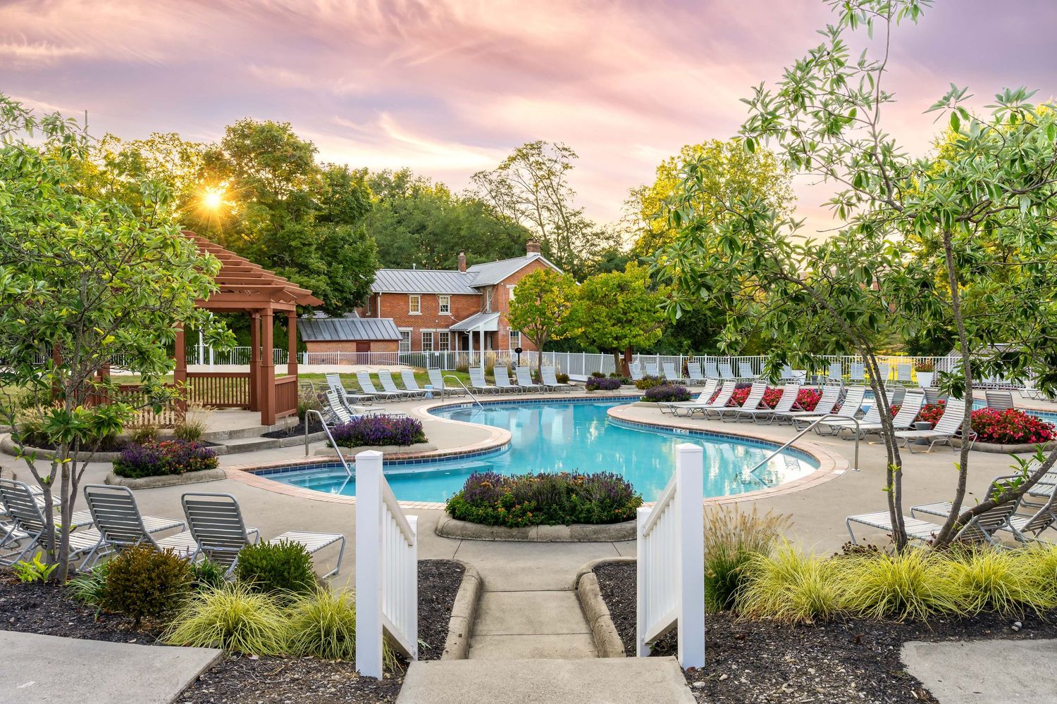 Luxury multi-family apartment poolside area with lounge chairs and gazebo at sunset surrounded by lush landscaping.