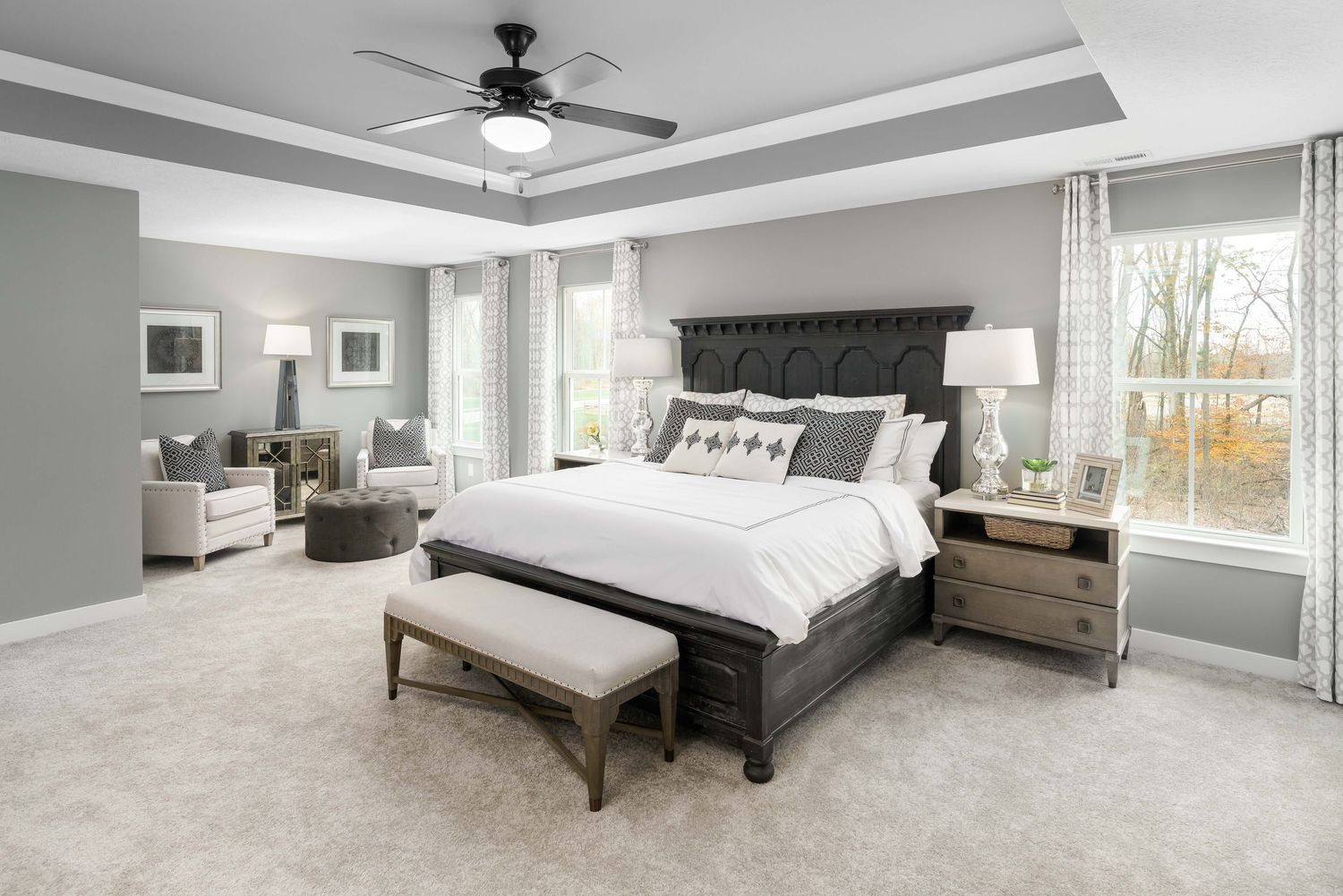 Model home with spacious primary bedroom with tray ceiling, plush carpet, dark wood bed frame and large windows overlooking backyard.