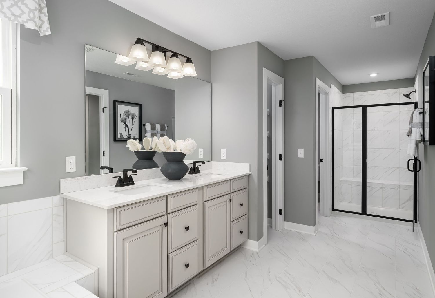Model home grey bathroom interior with white vanity, framed mirror, glass shower door and recessed lighting.