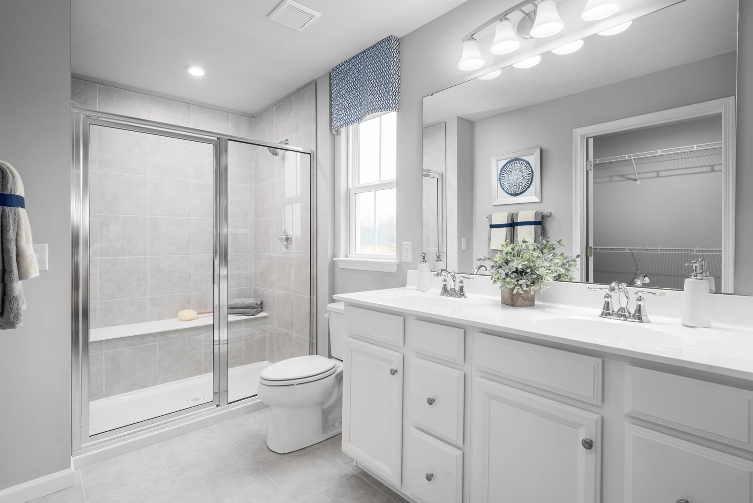 Model home white bathroom with glass shower enclosure, double vanity, and gray walls features bright recessed lighting.