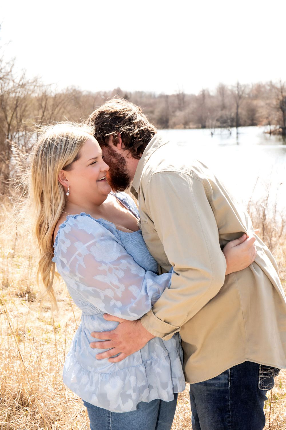A tender moment shared between a couple by a winter lake with dried grasses in the background.