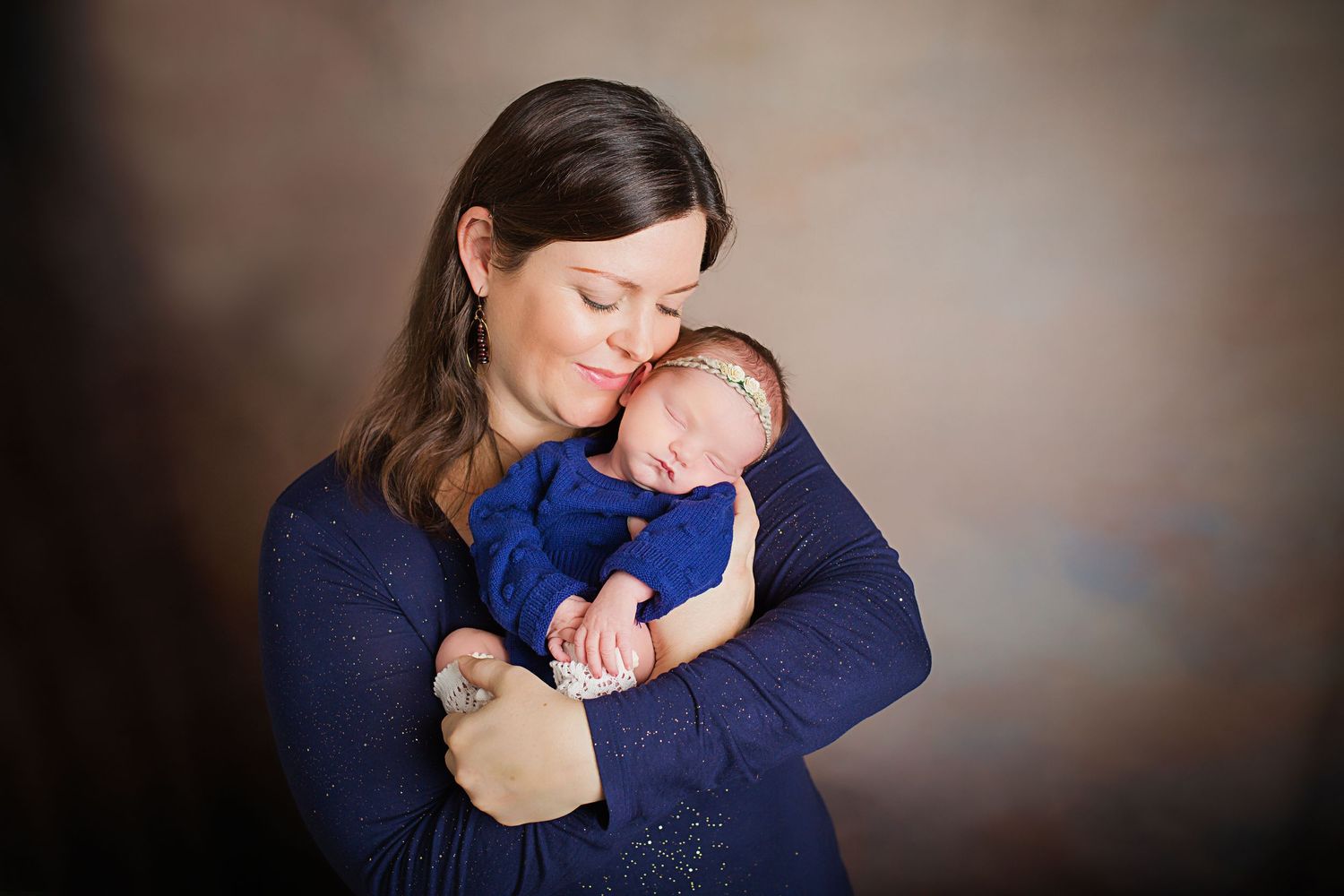 A new baby in navy sleeper is nestled against a mother wearing a dark blue sweater.