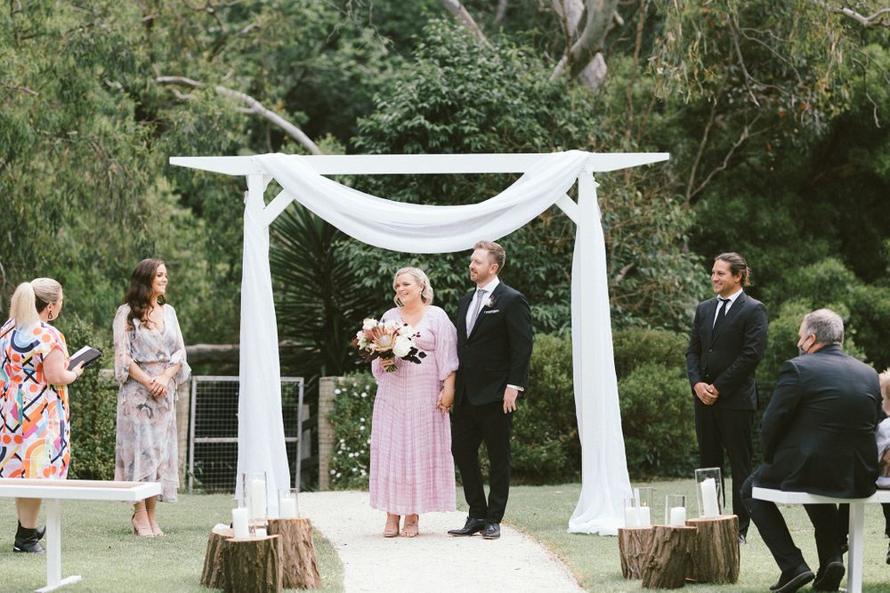 Outdoor wedding ceremony setup with white fabric draped arch and pink floral decorations.