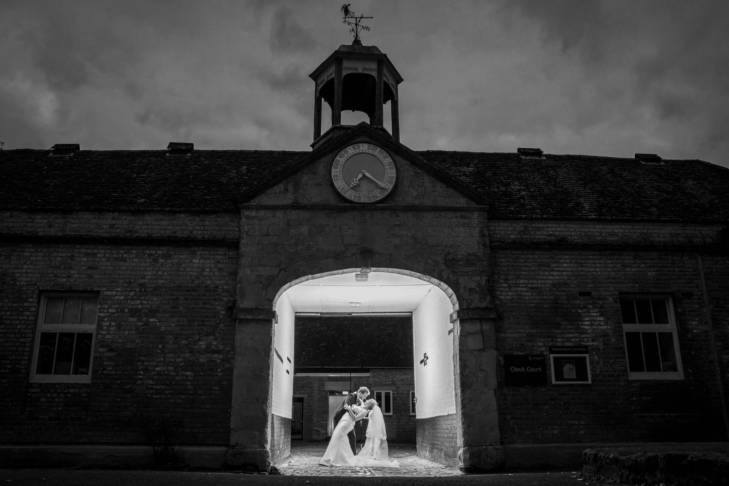 Black and white wedding photo of couple kissing under arched brick building with clock tower at night.