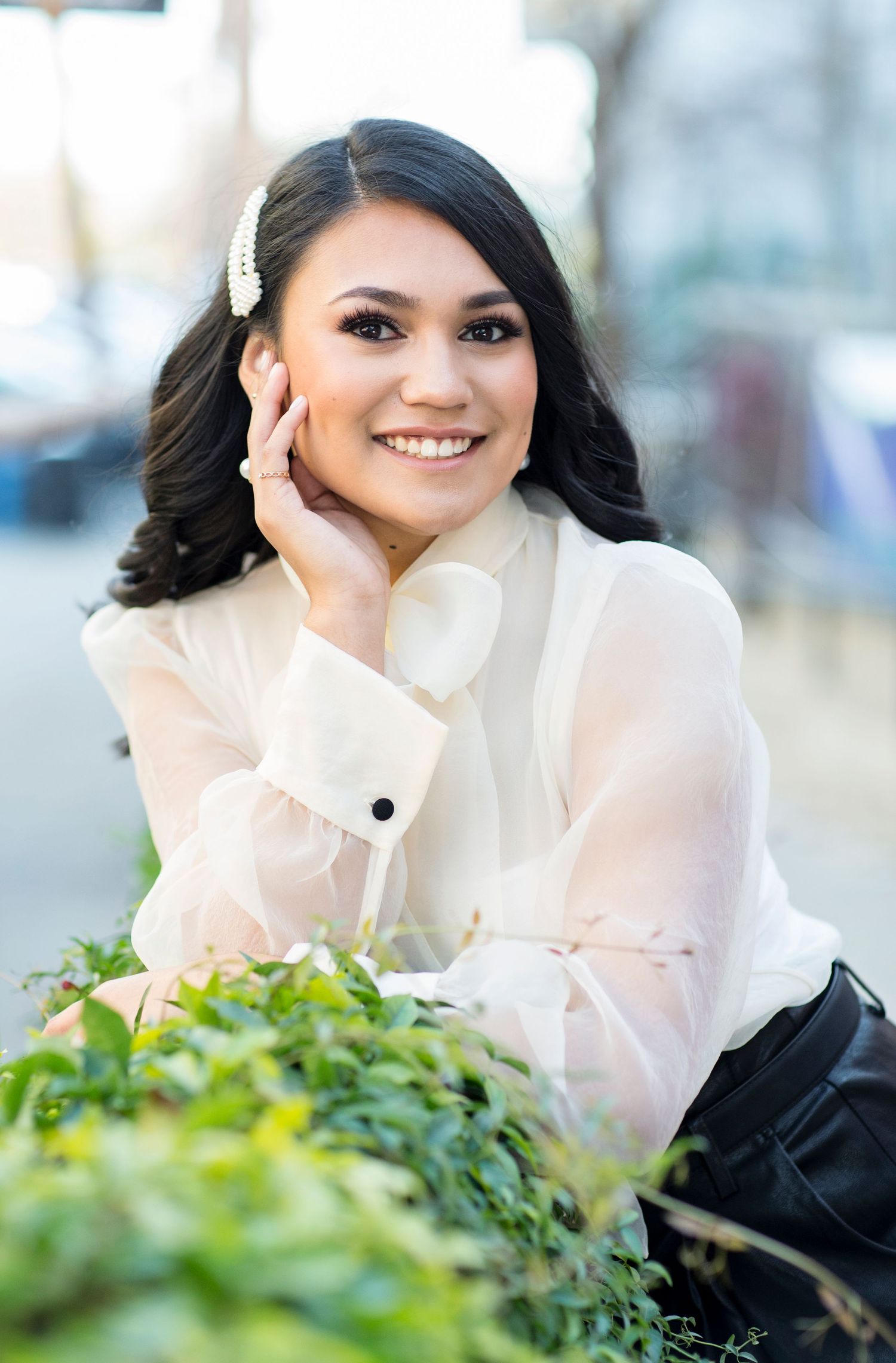 Portrait of someone in a sheer white blouse smiling warmly while posing outdoors near green foliage.