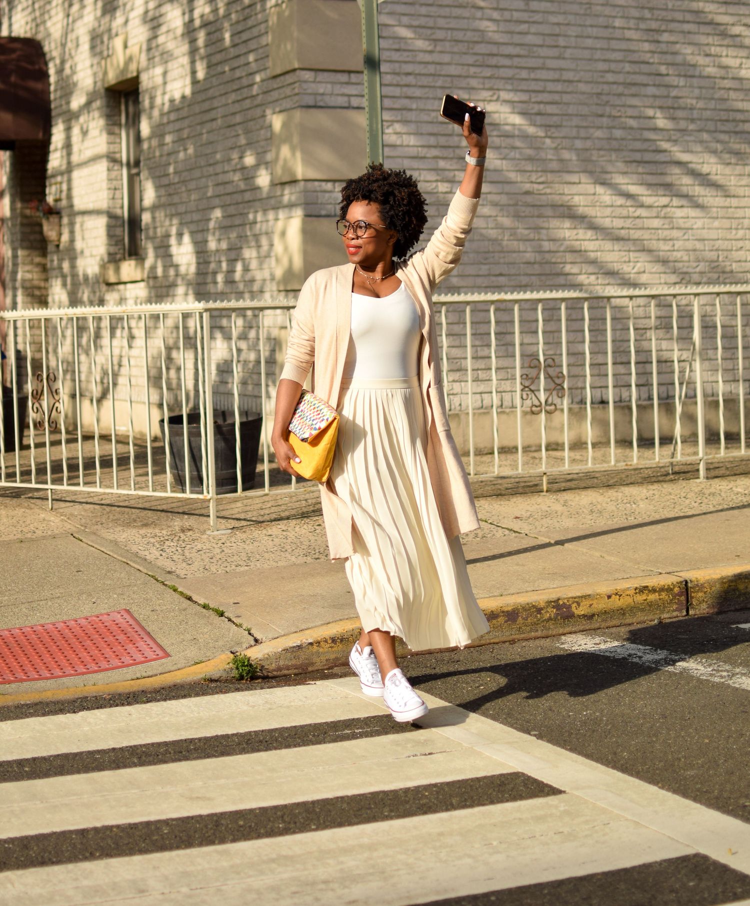 Stylish woman in flowing white dress crossing Hoboken, New Jersey street while holding phone and yellow purse in urban setting.