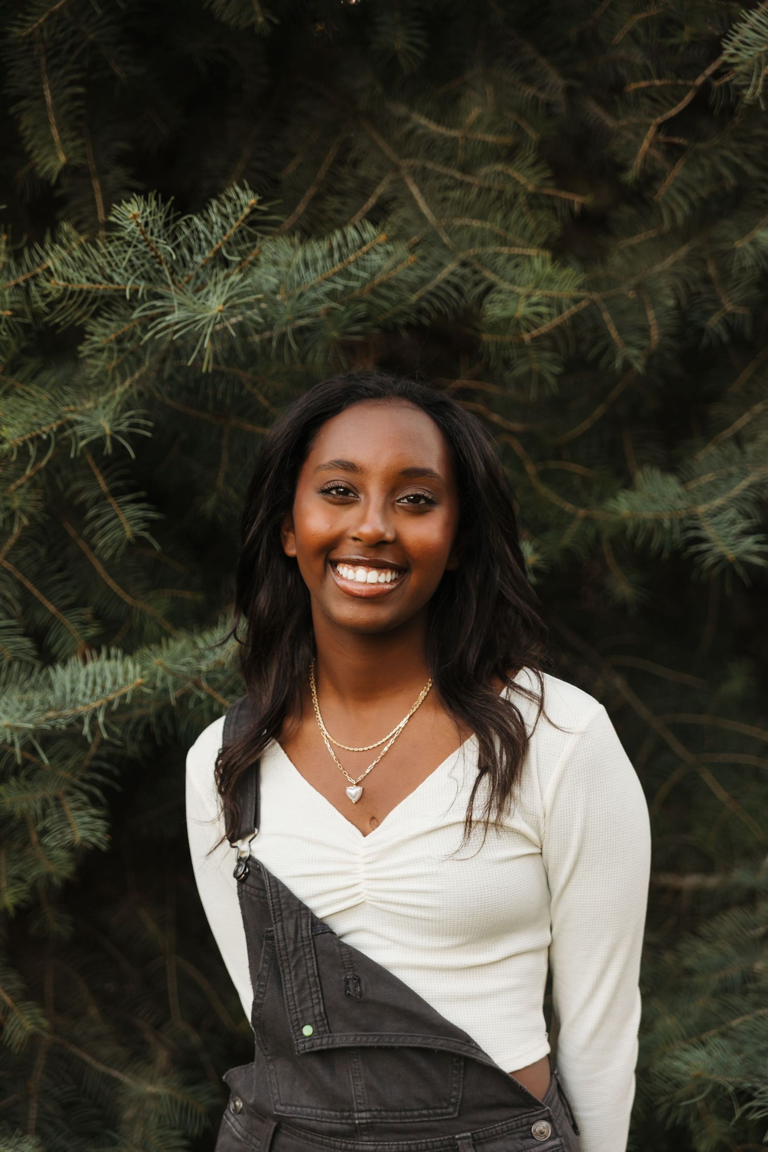 Portrait against evergreen trees of someone in white top and dark overalls with a bright smile.