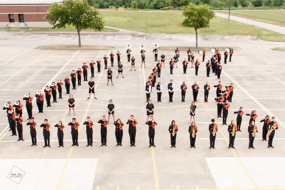 Marching band members form letters on pavement in orange and black uniforms.