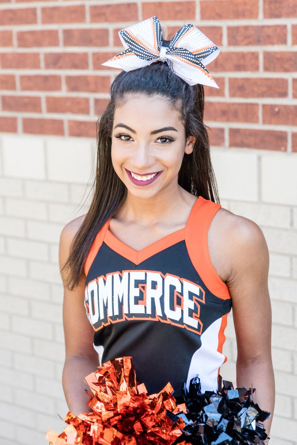 Cheerleader in black and orange Commerce uniform with bow poses against brick wall.