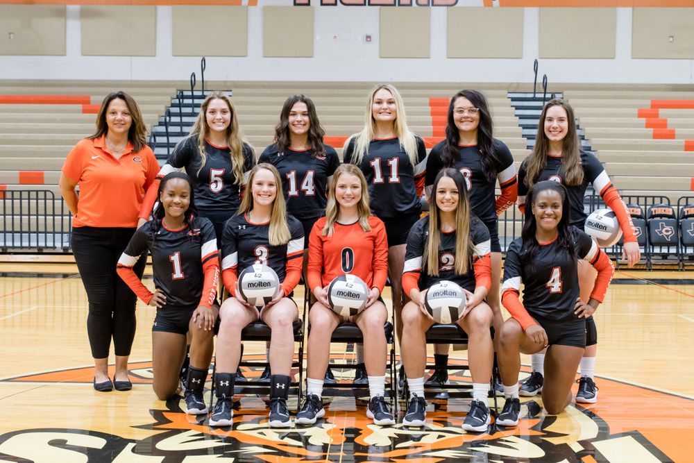 Volleyball team poses together in black and orange uniforms holding volleyballs.