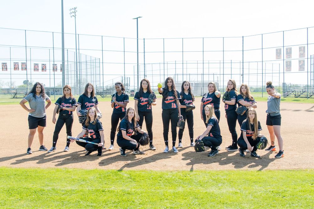 Softball team poses together on field with bats and gloves in matching dark uniforms.