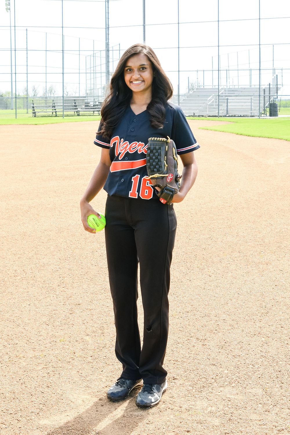Softball player in navy Tigers uniform holding glove and ball on dirt field.