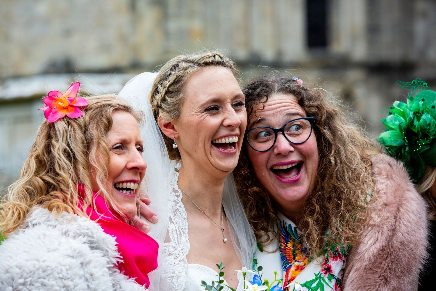 Three wedding guests share laughter and joy outside a stone church.