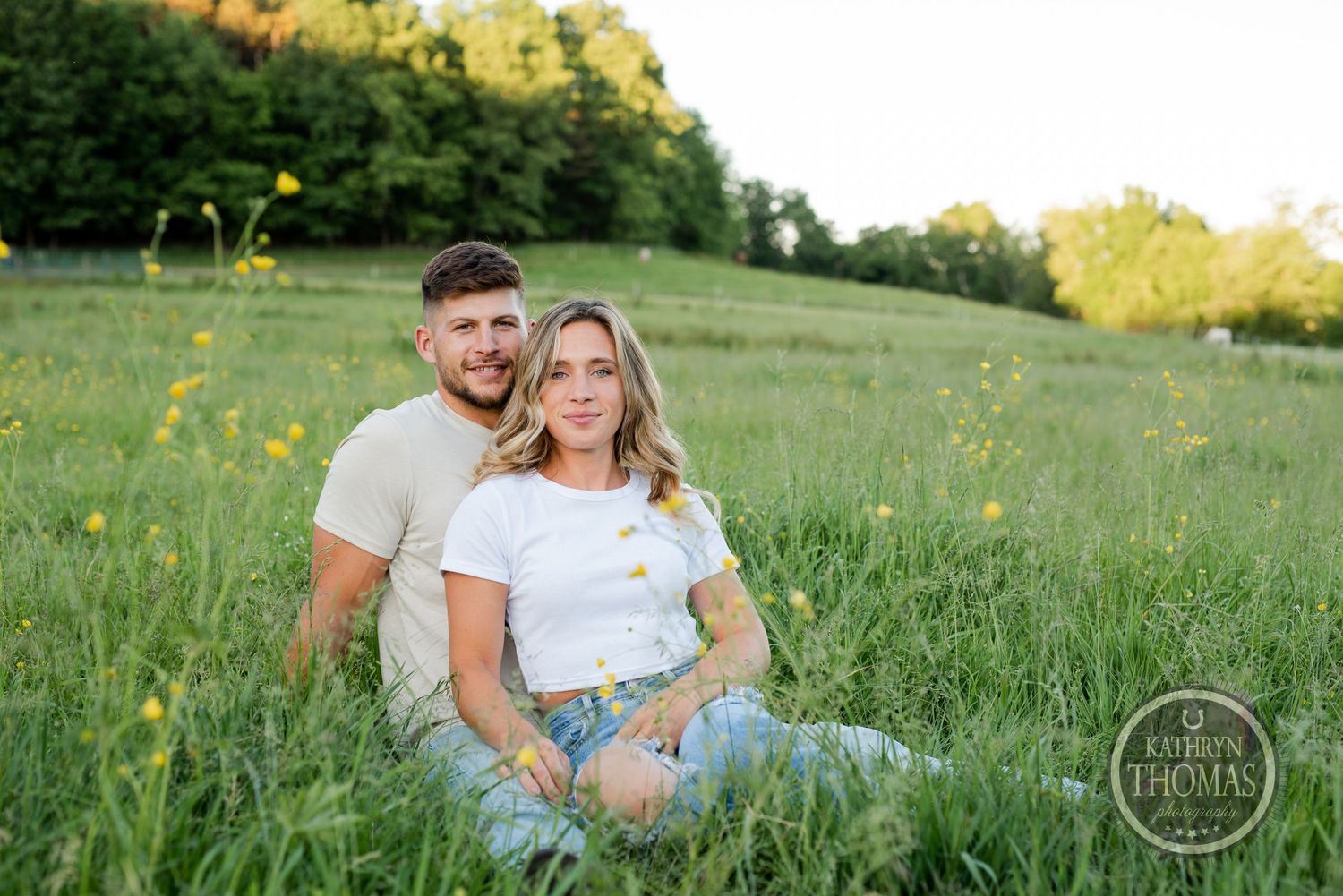 Couple in matching white t-shirts sitting together on grassy hillside with trees in background.