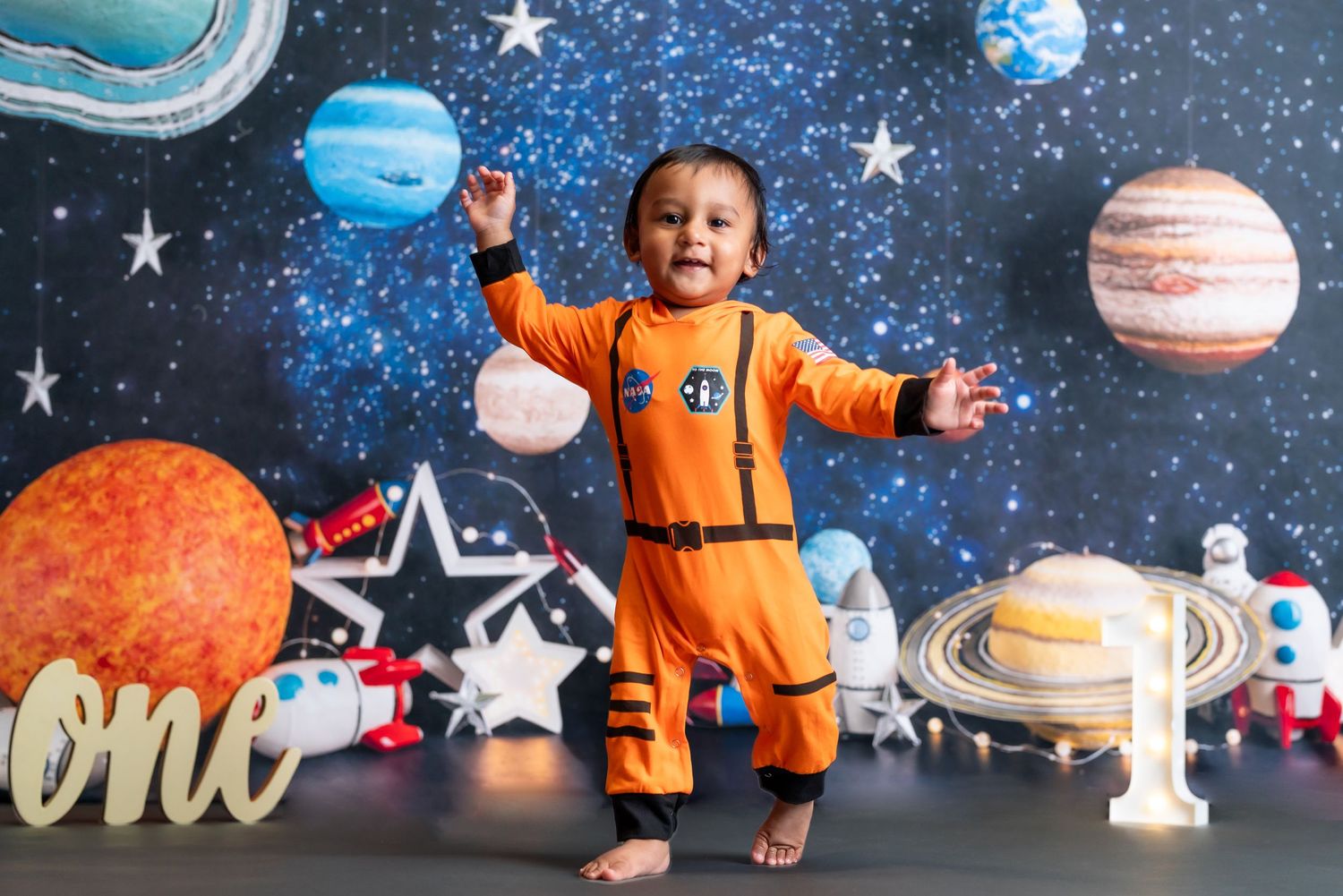 Baby in orange astronaut costume standing against space-themed backdrop with planets.