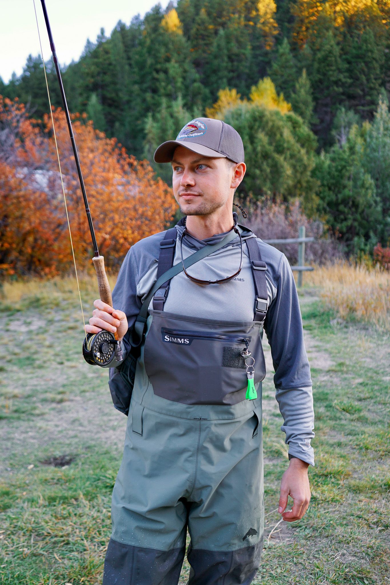 Fly fisher in waders and baseball cap holding rod with autumn foliage and forest in background.