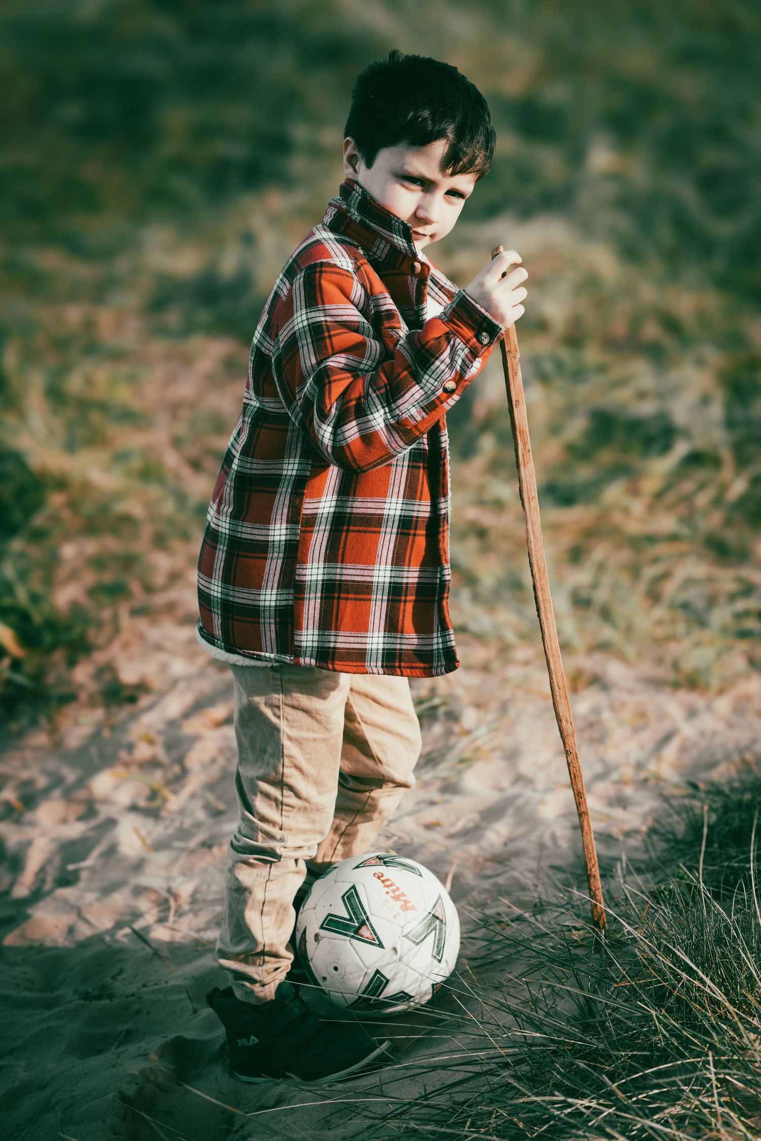 Young person in plaid shirt standing with walking stick and soccer ball in rustic outdoor setting.