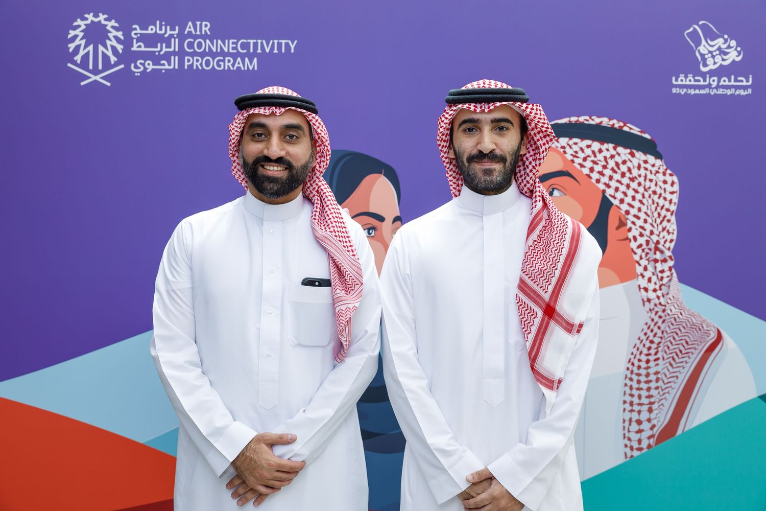 Two men in traditional white thobes and red and white checkered headdresses pose together.
