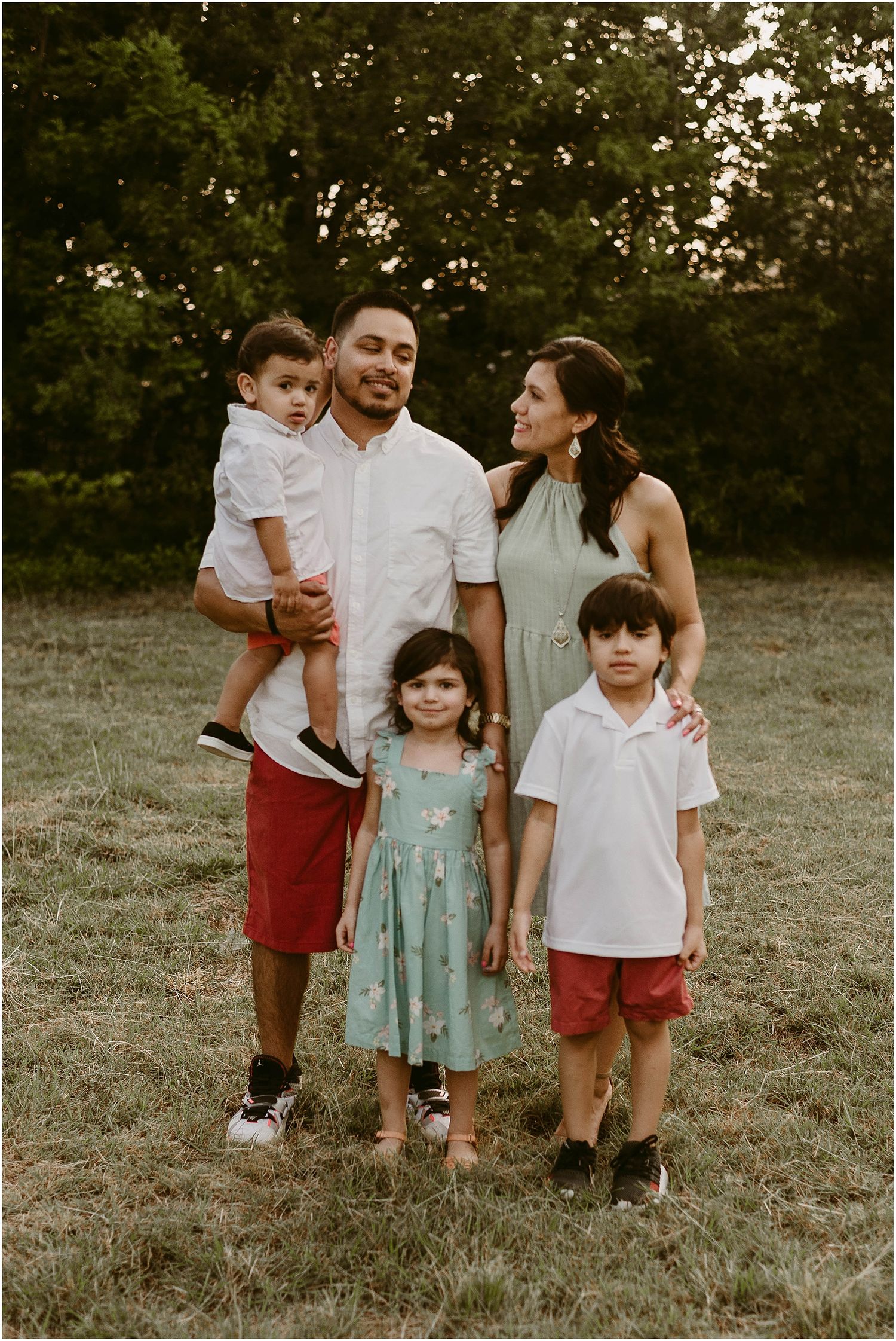 A loving family of five poses together outdoors in coordinated red and white outfits on a grassy field.