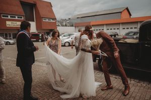 Wedding party helps with bride's dress in front of vintage car on cobblestone street.