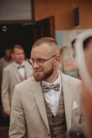 Wedding guest in beige suit and bow tie smiling at outdoor venue.