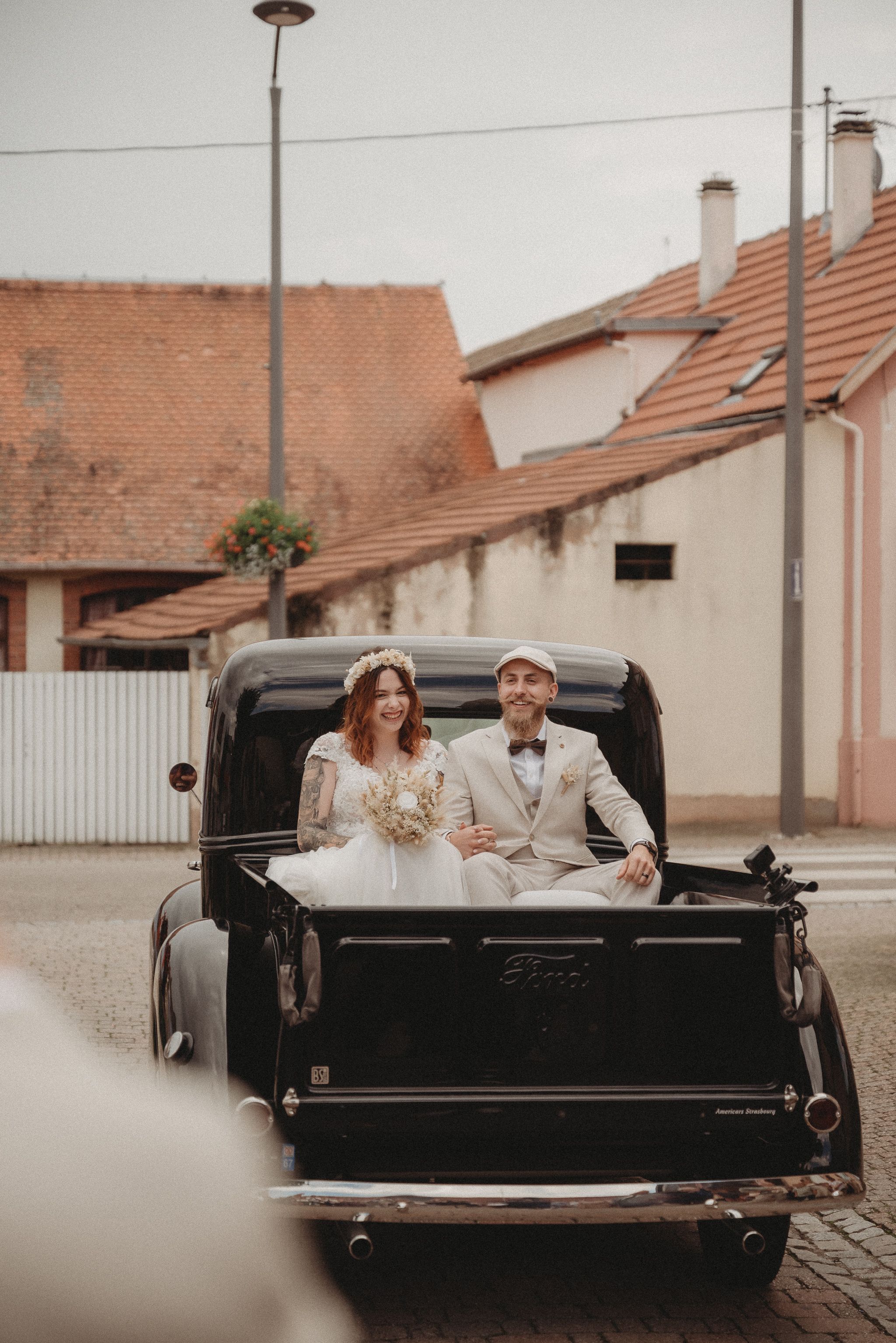 Couple in wedding attire pose in the back of a vintage Ford truck on a cobblestone street.
