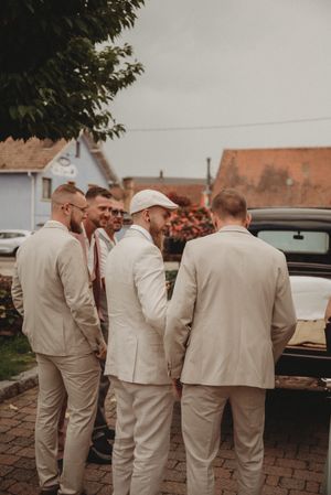 Groomsmen in cream-colored suits gather outside near parked cars.
