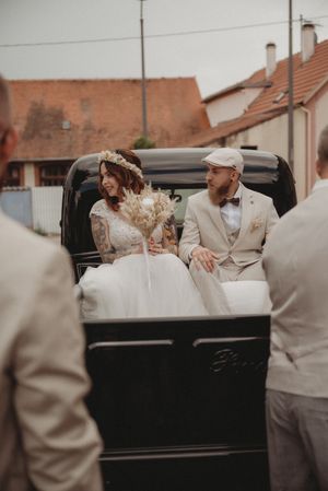 Newlyweds seated in a classic black vintage car with wedding guests surrounding them.
