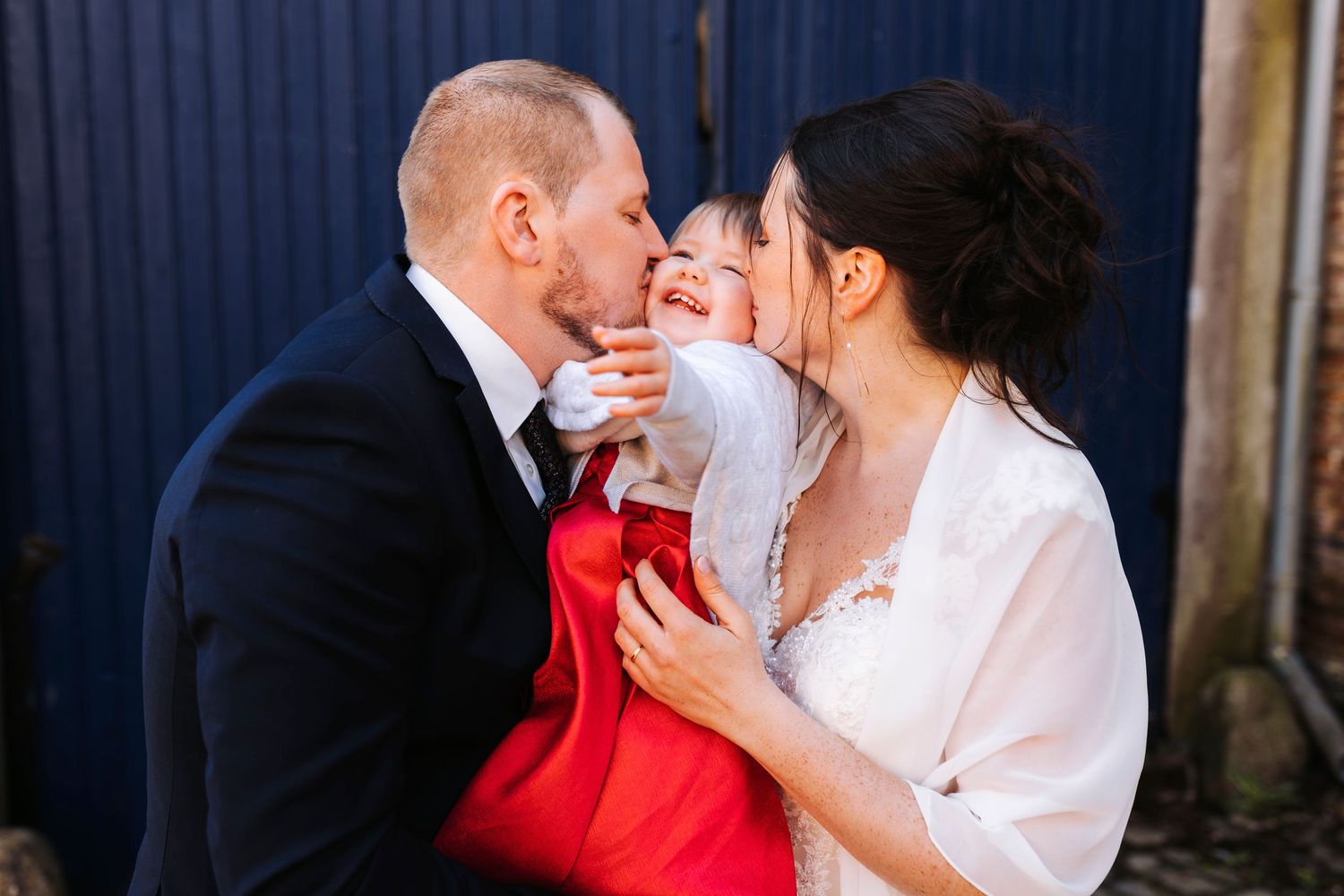 Romantic couple shares an intimate moment against a blue wall with the bride wearing a white top and red accent.